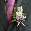 A close-up image shows the upper torso of a man dressed for a wedding in New Barnet, his attire captured in soft natural light as if he is standing just outside a church or registry office. He wears a dark charcoal grey suit jacket with a fine, almost brushed texture visible along the lapel and sleeve, over a crisp, pale dress shirt patterned with a subtle check that peeks out at the collar and cuffs. At the centre of his look is a vivid magenta silk tie, neatly knotted and gleaming slightly, its rich colour suggesting confident celebration. Pinned carefully to his left lapel is a handcrafted buttonhole, the true focal point of the scene. Slender, unopened cream and white buds rise at the top of the arrangement, reminiscent of snapdragons or lisianthus just about to open. Beneath them, soft pink and gentle lavender-purple blooms cluster together, like sweet peas or stock, their petals cradling the light and giving a cloud-like softness. Tiny white sprigs of gypsophila thread through the flowers, creating a misty, romantic haze, while a few deeper purple florets add depth and contrast. At the base, glossy dark green leaves anchor the design, interwoven with feathery, brighter green foliage that arcs lightly outward, giving movement and airiness. A delicate lavender satin ribbon ties around the stems in a precise bow, echoing the lilac tones of the petals and suggesting the careful hands of a local florist. The whole composition feels quietly joyful, a refined boutonniere perfectly suited to a spring or summer wedding in New Barnet, perhaps just moments before the groom joins guests gathering near local venues and leafy parks.