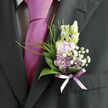 This image shows a refined boutonnière resting against a dark, formal jacket, a tiny floral artwork that seems to bring the soft light of a New Barnet morning into focus. At its heart, lilac flowers open in gentle layers, their petals cool and smooth, brushing softly against buds of creamy white. Threaded between them are clusters of airy white baby's breath, so fine they resemble a veil of mist drifting across a spring meadow. Surrounding the blooms, a collar of deep green leaves glows with a subtle, natural shine, their surfaces catching the light in graceful curves that echo the line of the lapel. Slim strands of green grass reach upwards, giving the piece a slightly untamed, garden-fresh feel, as though it were gathered near Hadley Common and then shaped by a careful, professional hand. The stems are neatly bound with a sheer lavender ribbon, tied flat and secure, its soft sheen linking the florals to the nearby tie and shirt. The colours-lilac, white, and rich green-stand out beautifully against the charcoal suit fabric, suggesting romance without being overly showy. You can almost imagine the faint perfume: a whisper of blossoms, clean and sweet, mingling with the crisp scent of freshly cut stems. Designed for weddings, proms, or black-tie events across New Barnet and the surrounding lanes, this boutonnière represents the quiet expertise of a local florist who understands how to balance elegance, comfort, and personal style in a single, perfectly proportioned piece.
