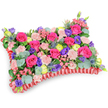 Seen from a gently elevated, oblique angle against a clean white background, this pillow-shaped floral tribute reads like a soft, comforting embrace. The rectangular cushion is densely packed with fresh blooms: a chorus of hot pink roses - some tightly furled, others beginning to unfurl into velvety spirals - creates the dominant, warm heartbeat of the design. Scattered among them are pale shell-pink carnations whose frilled edges catch the light and add a tactile, ruffled softness. Deeper notes of purple lisianthus (eustoma) punctuate the composition with bell-shaped depth, while cheerful lemon-yellow ranunculus offer sunny, buttercup-like accents that lift the overall palette. Cool, architectural silver-blue eryngium thistles introduce a spiky, wild texture that contrasts beautifully with the rounder blooms, and rounded eucalyptus leaves in silvery green weave through the surface, adding a fresh, aromatic veil of foliage and subtle movement. The whole arrangement is set within a deep red, pleated satin ribbon border that forms a ruffled frame around the cushion, defining its classic memorial silhouette and lending an elegant, almost textile-like finish. Soft, natural light reveals the layered construction - tight underlayers giving structured support beneath the airy surface - and the scent is quietly floral, dominated by rose and eucalyptus. Traditionally used as a sympathy pillow or memorial cushion, the design speaks more of celebration and remembered warmth than solemnity; its vibrant fuchsia, violet, lemon and blush tones convey affection and enduring memory. A local florist's hand is evident in the careful clustering and balanced contrasts, the kind of work you might see sent to a service near New Barnet station or placed with care in Oak Hill Park remembrance settings.