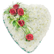 The photograph shows a refined heart-shaped funeral tribute placed against a plain white background, allowing every flower and ribbon edge to be clearly seen. The heart is fully covered with tiny, tightly packed white chrysanthemums, creating a smooth, pillowy surface that appears soft to the touch and symbolises purity and peace. At the upper left curve of the heart, three deep red roses form a striking cluster, their petals velvety and beautifully layered, standing out boldly against the white chrysanthemum base. Around them, glossy green leaves, wispy fern-like foliage, and a scattering of small white filler flowers add texture and a natural, garden-like feel. From this cluster, thin strands of bright green ornamental grass sweep diagonally across the heart, gently guiding the eye down towards two more individual red roses placed further to the right and slightly lower on the design. This subtle diagonal of red blooms and green lines adds rhythm and movement, as though the roses are quietly drifting over a bed of snow. The perimeter of the heart is carefully finished with a rich dark green satin ribbon, neatly folded to trace the curves and provide a clear, elegant border. The combination of crisp white, deep red, and varying shades of green creates a dignified, traditional tribute that many families in New Barnet might choose for a funeral, memorial service, or graveside visit. The arrangement feels calm, respectful, and full of love, offering a gentle way to say goodbye and to honour cherished memories.