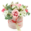 At the centre of the image, a beautifully composed hatbox bouquet rests against a spotless white background, creating the impression of a carefully staged studio photograph. The round, cylindrical box is a soft, pastel pink with a velvety matte surface, wrapped with a slim coral-red satin ribbon that catches the light with a subtle sheen. The ribbon is tied into a neat, modest bow on the front, where a cream-coloured rectangular tag with green writing hangs delicately, as if waiting to carry a few heartfelt words before being delivered to a doorstep in New Barnet. From the open top of the box, a luxurious flourish of flowers rises in a rounded, dome-like shape, full and generous but never heavy. A standout feature is a large, fully opened pale pink rose whose petals deepen in colour toward the centre, forming a swirling heart that draws the eye. Around it cluster white daisy-style chrysanthemums with vivid green centres, their simple, open faces bringing a bright, friendly feel that might remind someone of fresh greenery along local streets and gardens. Slender white alstroemeria, their petals finely marked inside, weave through the arrangement with gentle elegance. Deeper pink blossoms-likely spray roses or lisianthus-sit in smaller clusters, introducing pockets of rich, romantic colour. Silvery-green eucalyptus leaves spill slightly beyond the mass of blooms, their rounded shapes and muted tones adding a cool, aromatic freshness, while darker foliage and small budding stems quietly support the design. The overall impression is of a thoughtful, modern floral gift-perfect for a birthday, a romantic gesture, or a quiet thank-you-crafted with the kind of attention to detail one would expect from an experienced local florist serving the New Barnet community.