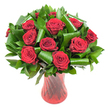 A lush, rounded bouquet of a dozen or more deep crimson roses fills the frame, each bloom opened fully to reveal tight, velvety spirals of petals that catch the light like soft, wine-coloured silk. The rich, dark red of each head is intensified by gentle, even illumination that reveals the delicate veining and layered texture of the petals, while a classic rose scent seems to lift from the cluster. The roses are generously framed and supported by an abundance of dark green foliage: flat, glossy leaves that form a dense verdant backdrop (suggestive of Salal or Ruscus) and broader, emerald leaves-shaped and rolled into elegant curls and tubular loops-whose architectural folds create graceful arcs around the blooms. These sculpted leaves add a modern, structured counterpoint to the romantic softness of the roses, lending both depth and movement to the composition.

The arrangement sits in a tall, translucent ruby-red glass vase with a subtle ribbed or twisted surface that catches vertical highlights and echoes the passionate hues of the flowers. The vase's tapered silhouette and faintly flared rim give the display a refined, gallery-like presence against a clean white background, isolating the colours and textures so they read purely and powerfully. As a local florist might say, this is the sort of bouquet we craft for anniversaries, Valentine's moments, or as a refined centrepiece for an intimate dinner. Freshly arranged and ideal for delivery across New Barnet and the surrounding neighbourhoods, it conveys classic romance, quiet confidence, and the tactile pleasure of well-handled blooms.