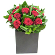 A compact, contemporary bouquet where deep crimson roses gather like a chorus at the center, each bloom velvety and perfectly spiraled, commanding attention without excess. Broad, glossy green leaves have been carefully curled and layered around the roses to create a rhythmic shell of texture and movement; their waxy sheen catches the light and gives the composition architectural clarity. The blooms sit low and confident in a matte black square vase whose geometric simplicity grounds the display and heightens the lavish red and emerald contrast. Lighting is bright and steady, as if photographed in a clean studio near New Barnet Station, catching the satin highlights on each petal and the subtle reflections on the foliage. If you lean in, the scent is a delicate, classic rose perfume moderated by cool, green notes-freshly cut leaves and a faint, almost tropical crispness from the broad greenery. Every detail feels intentional: the roses read as declarations of affection while the sculpted leaves lend restraint and modernity, making the floral statement equally appropriate by a candlelit anniversary table or on the reception desk of a contemporary office along Station Road. As a local florist familiar with Brunswick Park and the quiet streets off the Great North Road, I value how this design transforms a room without overwhelming it, offering a poised combination of romance and urban style. The tactile contrast between silky petals and firm foliage invites touch, and the whole arrangement presents as a thoughtful, sophisticated gift from a neighbourhood florist who understands New Barnet's quiet elegance.