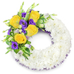 A solemn, lovingly crafted circular wreath, photographed against a soft white studio background, designed to read as the letter 'O' for a memorial tribute. The main body of the wreath is formed from hundreds of small, pristine white spray chrysanthemums-tightly clustered to create a cloud-like, velvety surface that reads as a single, reverent field of white. The petals are close and layered, producing a soft tactile texture that suggests quiet and unity. Nestled on the upper left curve is a deliberate, hand-tied spray of colour: several fully opened yellow roses, their butter-to-gold petals gently unfurling, paired with deep royal-purple lisianthus (Eustoma) whose ruffled, cup-shaped blossoms add a sumptuous, cool contrast. A few unopened lisianthus buds in pale lavender and lime-green peek through, lending depth and the promise of unfolding life. Interspersed throughout the spray are glossy, rounded leaves-some with subtle variegation-and delicate fern fronds that give the cluster a natural, garden-like silhouette and a restful green frame. A slender, dark-purple crinoline or mesh ribbon peeks from beneath the white chrysanthemum ring at both inner and outer edges, a discreet echo of the purple blooms that ties the composition together. The open centre emphasizes the traditional wreath form while the subtle scent of roses and green foliage would register as faint and comforting. This arrangement reads as an expression of peaceful farewell-respectful, handcrafted, and suited for funerals or memorial services across New Barnet and nearby Brunswick Park, offered with the quiet care of a local florist experienced in sympathy tributes.