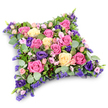 A tender floral cushion tribute, shaped like a soft rectangular pillow with curved corners, is shown resting against a bright white background, the simplicity of the setting allowing the colours and textures to radiate gently. The surface of the cushion is densely covered with fresh flowers, so full that they form a seamless layer of petals. At the forefront are large roses in shades of vivid fuchsia, rosy pink, and creamy light yellow, many in full bloom, their open centres revealing spirals of velvety petals that catch the soft light. Between these classic roses, deep purple lisianthus add a slightly cooler, more contemplative tone, their layered, ruffled petals contributing to the arrangement's rich, tapestry-like feel. Sprinkled among the larger flowers are smaller blush spray roses and fine clusters of lilac and lavender-toned filler flowers, similar to statice, which lend a delicate, feathery texture and fill even the tiniest gaps. Around the edges and nestled within the design, a subtle mix of greenery supports the composition: rounded, silvery eucalyptus leaves provide a calm, misty green, while finer, darker foliage adds gentle structure. The mood of the piece is one of quiet compassion and thoughtful care, echoing the peaceful atmosphere of New Barnet's residential streets and nearby green spaces. Ideal as a funeral tribute, a memorial arrangement, or a grave-side cushion delivered locally, this design expresses love, gratitude, and a wish for serene rest, offering families in New Barnet a comforting floral gesture at times of loss.