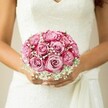 There's a gentle magic in this New Barnet bridal scene: vibrant roses, their petals like dawn skies, swirl in shades from hushed blush at the centre to exuberant magenta at the tips. Each flower is expertly nestled by a florist, creating a dense, round bouquet brimming with texture and light. Wisps of snow-white baby's breath interlace among the blooms, their tiny blossoms lending a veil-like softness that diffuses the intense colour, much like mist lifts over Oak Hill Park at sunrise. The bouquet is lovingly cradled just beneath the bodice of a white lace wedding gown, the intricate floral pattern echoing the blooms it frames. A subtle, pale manicure catches the light as the bride's hands warm the fresh stems, and a delicate sash ribbon trails beside her wrists, adding to the air of anticipation. The neutral, softly blurred background serves to heighten the impact of the bouquet's artistry, suggestive of those quiet, breath-held moments before vows are spoken. This image captures the scent, softness, and shimmer of a day built on hope and love - an enduring token of a New Barnet ceremony, and a masterwork of local floral storytelling.
