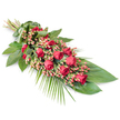 A refined, elongated sympathy spray featuring deep red roses and soft berries is laid diagonally across a clean white background, its poised form suggesting a final, gentle gesture of farewell. The upper left shows the stems drawn together and wrapped securely in a broad green leaf band, offering a polished, formal finish, as if prepared for presentation at a funeral service in a New Barnet church or chapel. From this neat binding, the arrangement fans out into a long, tapering design that widens gracefully through the centre before narrowing to a delicate point at the lower right. Throughout the length of the spray, rich, velvety red roses take centre stage. Some are tight buds with petals still cupped, others are just beginning to unfold, and a few are fully open, revealing deep, layered hearts that speak of enduring love, respect, and remembrance. Threaded between them are clusters of small, rounded Hypericum berries in warm pinkish-red tones, their slightly lighter hue and smooth texture providing a subtle contrast to the lush rose petals. Beneath and around the flowers, a bed of dark green foliage creates structure and depth: broad, glossy leaves form a strong base layer, while slender palm fronds and finer sprays of greenery extend beyond the main outline, suggesting gentle movement like a breeze along nearby Oakhill Park. The bright white background keeps the focus firmly on this traditional funeral tribute, emphasising the interplay of reds and greens. It feels quietly solemn yet comforting, a thoughtfully arranged floral expression of sympathy and support for families in and around New Barnet saying their final goodbyes.