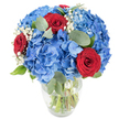 A richly textured bouquet sits in a clear glass vase, photographed against a soft white backdrop so each colour and form reads clearly. The dominant notes are voluminous clusters of sky-blue hydrangea, their many small petals overlapping to create a plush, cloud-like mass of cool, cobalt-tinted blue that feels both breezy and substantial. Punctuating that expanse are deeply saturated red roses, their velvet petals tightly curved into classic spirals that read as intimate, passionate accents against the cool blue. Scattered between the larger blooms are airy clouds of white baby's breath, tiny star-like florets that lend a delicate, whispery softness to the silhouette and ease the contrast between the strong colours. Muted, rounded eucalyptus leaves and fresh green foliage weave through the stems, their soft sage tones and smooth surfaces adding an herbaceous, calming note and a subtle, clean fragrance when held close. The transparent vase has a gently rounded base that tapers slightly to the rim, and the green stems are visible through the water, a simple indicator of freshness and careful floral conditioning. Bright, even lighting emphasizes the saturated pigments and the tactile contrasts-the cool sheen of hydrangea petals, the velvety depth of roses, the feathery lightness of gypsophila. As a local florist would say, this arrangement feels celebratory and composed, suitable as a centerpiece for a dining table or as a thoughtful gift to mark anniversaries, birthdays, or moments of quiet devotion in homes around New Barnet and nearby Monken Hadley Common.