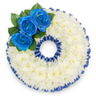 A bespoke funeral tribute fashioned in the precise shape of the number '0', photographed from above against a clean white background so every detail reads clearly. The foundation is a dense, pillowy field of small white spray chrysanthemums, their clustered florets forming a soft, tactile surface that calms the eye and suggests quiet consolation. Around both the outer rim and the hollow centre, a pleated royal blue satin ribbon has been carefully tacked in place, its ruffled scallops creating an elegant frame that echoes the saturated hue of the roses. At the upper left curve, a cluster of three vivid royal blue roses rises above the chrysanthemum bed, their petals unfurling with a velvety sheen and set against a few glossy, dark green leaves that add depth and a living accent. The blue may be natural or delicately enhanced to achieve that deep sapphire tone, a colour often chosen to reflect a loved one's service uniform, club colours, or a personal favourite. Soft studio light washes across the ruffled ribbon and the rounded chrysanthemum heads, casting gentle shadows that accentuate the wreath's sculpted form and solemn symmetry. This numerical memorial carries the same care and quiet dignity we bring to tributes delivered across New Barnet and neighbouring Brunswick Park-an arrangement meant to read as both personal marker and public act of remembrance. The overall mood is one of respectful reverence: clean, balanced, and considered, a floral language of peace and continuity for a service, wake, or grave-side farewell.
