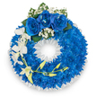 A solemn, meticulously arranged circular wreath photographed against a clear white backdrop: the main body is a plush, continuous ring of small, intensely vivid electric-blue chrysanthemums or similar small-petalled blooms, packed so closely they create a velvet-like, tactile surface. The deep blue tone is luminous, catching the light along each curled petal and giving the circle a sense of depth and quiet dignity. At the very top, three full-blown royal blue roses form a gentle focal point; their velvety petals unfurl in layered spirals and are softly haloed by tiny clusters of pure white baby's breath and a few dark green leaves that add botanical contrast. From the left side a graceful spray of pristine white orchids - likely Phalaenopsis with broad, open petals - arcs downward, introducing slender drama and a sense of fragile beauty; intertwined with the orchids is a delicate stem of pale yellow-green buds that suggest unfolding life and hope. The wreath's inner edge is finished with a crisp, ruffled white satin ribbon that frames the central opening and lends a formal, respectful border to the composition. The overall effect is one of refined sympathy and peaceful remembrance: colours that speak of calm, fidelity, and quiet reverence. Subtle floral scent and the soft play of studio light enhance the wreath's texture in the image. This is the kind of tribute chosen for funerals or memorials in New Barnet, suitable to place near New Barnet station, Brunswick Park, or local crematoria when families wish to combine dignity and floral care in moments of farewell.