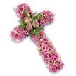 A richly detailed floral cross fills the frame, set against a bright white background that highlights its soft colours and intricate structure. The cross itself is entirely covered in a thick layer of blossoms, dominated by shades of blush pink, mauve, and light lavender, so that the flowers create a continuous, cushioned surface from top to base. At the central crossing point, three statement roses in a subdued dusty-rose and pale mauve tone are arranged together, their open petals curling back in elegant spirals. These roses are nestled among sprays of dark green leaves and fine fern fronds, which introduce depth and a hint of garden freshness. Stretching along the arms and down the length of the cross, clusters of lisianthus blooms form the main body of the design; their trumpet-shaped flowers, some with deeper coloured centers, add gentle waves of texture and tone. Scattered between them, smaller pink and purple accents and minute white flowers with soft yellow centers appear like tiny stars, catching the eye as you move across the arrangement. Along the edges of the cross you can just see glimpses of narrow deep-purple ribbon or groundwork, giving the design a polished finish. The overall effect is serene and deeply respectful, like a floral echo of the calm corners of New Barnet's residential streets and nearby green spaces. Crafted in the style of a dedicated local florist, this funeral cross would feel at home resting beside a coffin, on a memorial stand in a New Barnet chapel, or placed by the graveside, offering mourners a gentle symbol of faith, remembrance, and unspoken love.