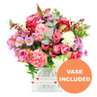 A generously full bouquet of mixed fresh flowers is displayed in a clear glass vase against a flawless white background, its colours radiating warmth and tenderness. The eye is immediately drawn to the cluster of pink roses at the centre, some in a saturated fuchsia tone and others in a gentle powder-pink, their layered petals opening outward in soft spirals. Surrounding them are ruffled carnations in shades of coral, peach, and blush, adding a feathery texture that makes the arrangement feel plush and inviting. Scattered among these larger blooms are small, daisy-like chrysanthemums in lavender and light purple, their vivid green centres offering a lively contrast, alongside creamy white flowers that bring calm balance to the composition. The blooms are encircled by abundant foliage: cool, rounded eucalyptus leaves with a silvery-blue tint mingle with darker green stems and occasional reddish-brown leaves, giving the arrangement depth and a slightly seasonal nuance, reminiscent of the varied greenery seen in New Barnet's local parks and garden fronts. All the stems are gathered into a modern, square-shaped glass vase with softly rounded edges, the base of which is wrapped in a neat white band decorated with repeating red hearts and the phrase all my love in flowing script, clearly signalling that this is a bouquet designed to express affection and gratitude. In the lower right corner of the photograph, an orange circular badge boldly states VASE INCLUDED in white capital letters, indicating that the flowers arrive ready to place straight on a table or windowsill, perfect for same-day delivery across New Barnet for birthdays, anniversaries, Mother's Day, or simply to brighten someone's day.