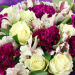 The image shows a close-up, thoughtfully composed bouquet that feels as if it has just been lifted from a New Barnet florist's workbench, its flowers gathered tightly together so every detail is visible. At its heart are several creamy white roses, some just beginning to open and others more fully unfurled, their petals silky and layered, with occasional hints of buttery yellow at the centres that add warmth and depth. Clustered closely among them are deep magenta and rich purple carnations, whose densely ruffled, almost feathery petals give the arrangement a plush, tactile richness, like soft fabric folded in waves. Threaded throughout, white alstroemeria bring an airy lightness; their narrow, trumpet-like petals are traced with fine pink and burgundy lines that create a sense of movement and artistic flair. Fresh green foliage, smooth and slightly glossy, weaves in and out, offering restful spaces of colour between the more dramatic blooms and shaping the bouquet into a rounded, abundant form. Around the outer edges, a gentle lavender wrapping can be seen, its muted, papery texture softly echoing the bouquet's purple tones and framing the flowers as a ready-to-give gift. The lighting is calm and even, drawing out the creaminess of the roses and the richness of the carnations, as though the bouquet were basking in natural light from a New Barnet shop window near the High Road. The overall mood is tender yet confident, making this arrangement well-suited for birthdays, thank-you gestures, or meaningful occasions shared with loved ones throughout New Barnet and surrounding areas.