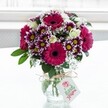A vibrant mixed bouquet sits proudly in a clear, subtly textured glass vase, placed against a softly glowing white backdrop that feels like daylight streaming through a New Barnet window. Deep fuchsia and magenta gerbera daisies, each with a velvety dark centre, form bold focal points that draw the eye first, their broad petals radiating outward like little suns. Around them, bicolour chrysanthemums in rich purple and white create a halo of intricate texture, their fringed petals tipped in plum and fading gently to cream, circling bright yellow centres that suggest warmth and optimism. Creamy white spray roses, some fully open and others still cupped in tight buds, are tucked thoughtfully between the larger blooms, adding a romantic softness and a hint of classic rose fragrance. Airy sprigs of white baby's breath weave through the arrangement like a fine mist, lightening the composition and giving it a dreamy, almost cloud-like quality. Deep green foliage anchors the flowers, its glossy leaves framing the colours and reinforcing the sense of freshly gathered stems. The vase itself is tied neatly at the neck with rustic twine, the natural fibres contrasting charmingly with the glass and suggesting a hand-finished touch, like something you might receive from a trusted local florist after a stroll along East Barnet Road. A small pale gift tag hangs from the twine, decorated with a delicate pink floral emblem and subtle lettering, hinting at a personal message for a birthday, thank-you, or just-because surprise. The overall impression is of joy and celebration captured in flowers, perfect for brightening a home near New Barnet station or marking a special moment shared with someone dear.