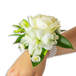 A delicate white wrist corsage sits gently on a fair-skinned wrist, photographed against a clean white background with soft, even studio lighting that highlights textures and subtle colour shifts. At the heart of the arrangement, a creamy ivory rose unfurls velvety petals in tight, elegant spirals; its soft sheen and powdery surface catch the light with a quiet glow. Nestled below and to the left, a larger white trumpet-shaped bloom - reminiscent of an Alstroemeria or a small lily - opens to reveal a pale green-yellow throat that adds depth and a whisper of spring freshness. Sprays of tiny, cloud-like Gypsophila drift around the main flowers, their fragile white dots creating a soft, airy halo, while a trio of small daisy-style florets with golden centers offers a cheerful, detailed counterpoint near the wrist. Several vivid green buds, some still closed, promise gentle movement and future bloom, and broad, glossy deep-green leaves frame the composition with a lush contrast that grounds the corsage. The floral cluster is secured on a chic multi-strand pearl bracelet: uniformly round, lustrous faux pearls loop snugly around the wrist, their cool, smooth texture balancing the blossoms' delicate softness. You can almost sense the faint, clean fragrance of freshly cut roses and green foliage. As a local florist would craft for brides, prom-goers, or gala attendees around New Barnet - from near the station to the edges of Monken Hadley Common - this piece reads as classic, refined, and quietly celebratory, designed to sit comfortably on the wrist while echoing timeless elegance in every petal and pearl.