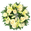 Viewed from above on a clean white surface, this circular sympathy wreath reads as a study in serenity: densely arranged creamy-white roses and soft, pale yellow trumpet lilies form a flawless ring, their tones balanced by a deep bed of fresh green foliage. About fifteen tightly furled roses create a plush, ivory core, each bloom opening into classic spirals that catch the light with a soft, powdery sheen. Between them, lemon-tinted lilies unfurl in various stages-some fully open with graceful, elongated petals and pale stamens, others poised as slender buds-offering a gentle rhythm of height and pause across the wreath's face. The greenery beneath is integral to the composition: feathery fern fronds weave through the flowers, adding airy, lace-like textures, while rounded eucalyptus-like sprigs introduce cool grey-greens and a calm, structured counterpoint. The overall effect is lush and intentionally full, the blooms tightly set so the eye moves in an unbroken circle, a quiet emblem of remembrance and continuity. There is a subtle sensory presence implied-the roses' soft, sweet perfume tempered by the lilies' fresher, almost citrusy note-yet the visual restraint keeps the tribute dignified and consoling. Crafted with careful layering and balance, this wreath would suit a funeral service or graveside tribute delivered with empathy to families in New Barnet and neighbouring Oakleigh Park, where thoughtful local floristry is often chosen to accompany moments of farewell. The composition's pale palette and verdant framing communicate peace, purity, and a comforting, steady grace.