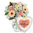 A joyful Mother's Day gift arrangement is pictured in bright, even light against a pure white background, making each soft pastel flower and shiny accent stand out. At the heart of the image is a full, carefully crafted bouquet displayed in a clear glass vase with a slightly rounded jar-style silhouette, its neck cinched with a loose bow of rustic twine that brings a relaxed, homely charm. The bouquet itself features several peach-pink roses at full bloom, their velvety petals spiralling open in generous layers, alongside bold peach gerbera daisies with rich dark centres that create a sunny, uplifting contrast. Nestled among these standout flowers are delicate clouds of white baby's breath, their countless tiny florets adding a gentle haze, and small white chrysanthemums with fresh pale green centres that offer extra texture and depth, much like the layered greenery found in New Barnet's local parks and leafy back gardens. Sprays of lush, mixed foliage in varied shades of green weave through the arrangement, enhancing the sense of movement and natural abundance. Tucked behind the bouquet, just visible above the rim of the vase and beside the balloon, is a small white card with softly coloured writing, hinting at a Happy Mother's Day message without fully revealing the words. To the right of the flowers, a large heart-shaped foil balloon is anchored discreetly to the arrangement. Its white surface is sprinkled with metallic gold and silver dots that catch the light, while a central peach heart holds the black inscription HAPPY Mother's DAY in a mix of bold print and flowing script. Together they create a tender, celebratory tribute, perfect for Mother's Day deliveries across New Barnet and the surrounding community.