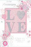 The photograph captures a single anniversary card presented as though ready to be given alongside a bouquet delivered across New Barnet. Its colour scheme rests in soft blush pinks, creamy whites and subtle greys, with delicate silver highlights that lend a refined, almost dreamlike quality. In the centre, the word LOVE stands out in embossed, glitter-kissed letters. The raised texture is clearly visible, suggesting a pleasant roughness beneath the fingertips, while the glitter itself shimmers gently rather than boldly, like moonlight on a calm evening walk through nearby Oakleigh Park. Surrounding this focal word, an intricate wreath of illustrated flowers, buds and leaves stretches across the card front. Each bloom is drawn with fine, flowing lines and shaded in pale pink and white, with hints of grey that add depth without overpowering the softness. The botanical shapes evoke the loose, romantic style of anniversary arrangements that might be crafted by a local florist, with slender vines looping and swirling against the dusky pink background. Small silver accents glint from petals and stems, giving the design a touch of luxury reminiscent of decorative foiling. The background itself is smooth and evenly lit, enhancing the contrast between the sleek card surface and the textured lettering. A thoughtful anniversary message in graceful script, though not fully visible, suggests heartfelt words of appreciation and enduring love. Overall, the card feels gentle, romantic and timeless, like a paper echo of an elegant floral display proudly placed in a New Barnet home.